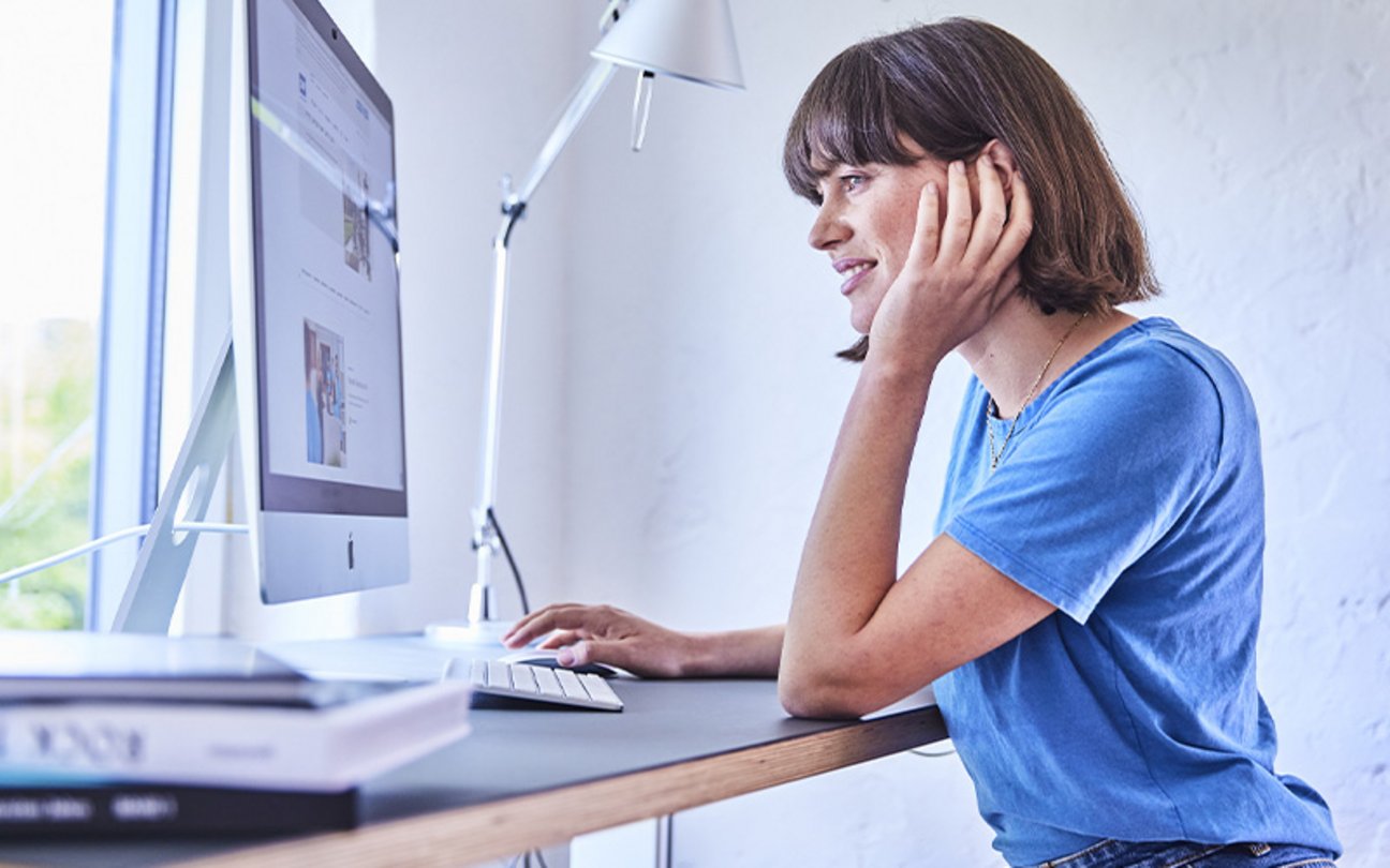 Junge Frau sitzt an einem Schreibtisch und arbeitet an einem Desktop-Computer in modernem, hellem Raum.