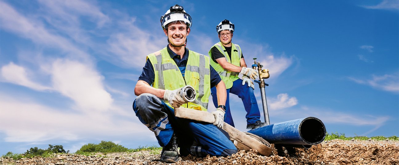 Zwei Bauarbeiter verlegen Wasserleitungen auf einer Baustelle unter blauem Himmel.