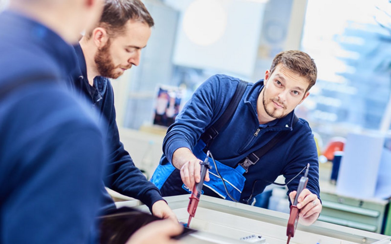 Das Bild zeigt einen jungen Mann, der mit technischen Messgeräten arbeitet und in die Kamera lächelt, neben ihm stehen zwei weitere Männer.
