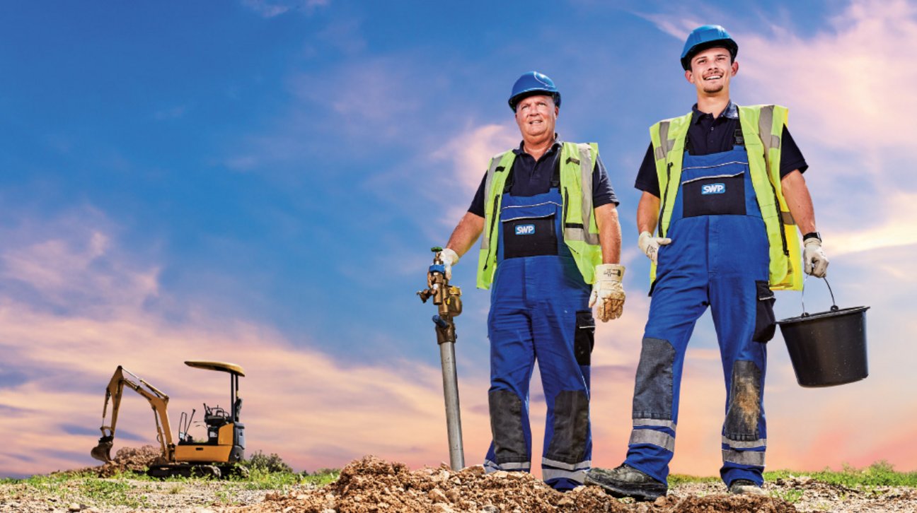 Detlef, Messstellenbetrieb und Moritz, Wasserversorgungstechniker Zwei Bauarbeiter mit Ventil und Eimer posieren auf einer Baustelle bei Sonnenuntergang.