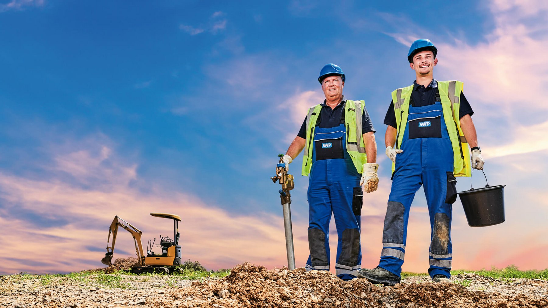 Zwei Bauarbeiter mit Ventil und Eimer posieren auf einer Baustelle bei Sonnenuntergang.