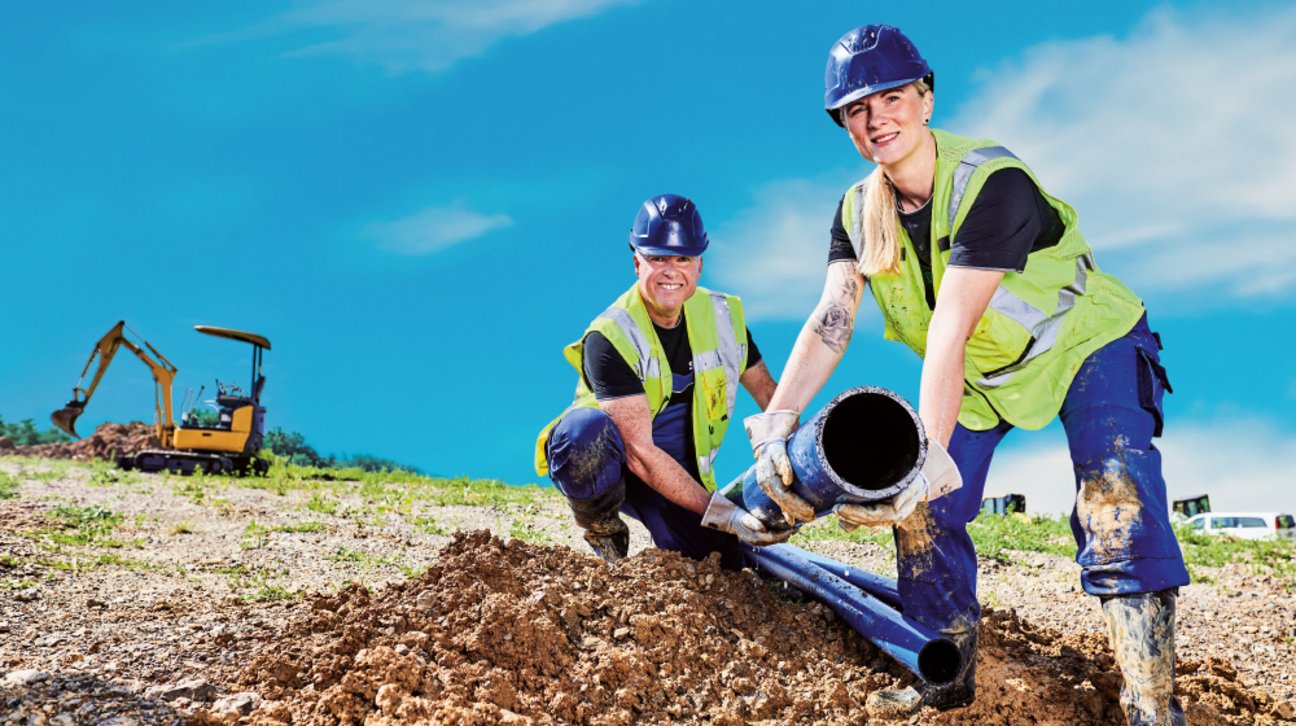 Andrea und Oliver, Team Energiedatenmanagement
Zwei Bauarbeiter verlegen blaue Rohre auf einer Baustelle mit Bagger im Hintergrund.
