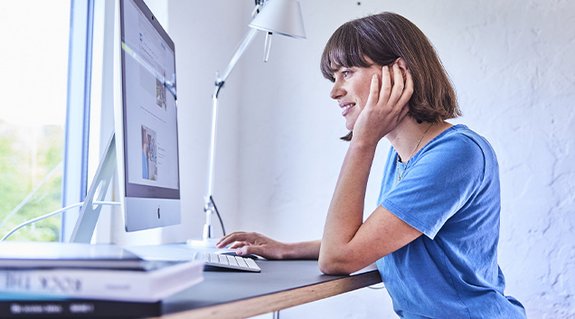 Junge Frau sitzt an einem Schreibtisch und arbeitet an einem Desktop-Computer in modernem, hellem Raum.