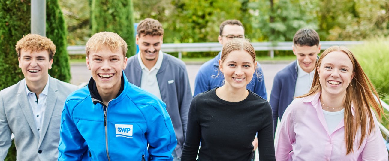 Eine Gruppe junger Menschen steht in einem Foyer, teilweise am Empfangstresen gelehnt und lächelt.