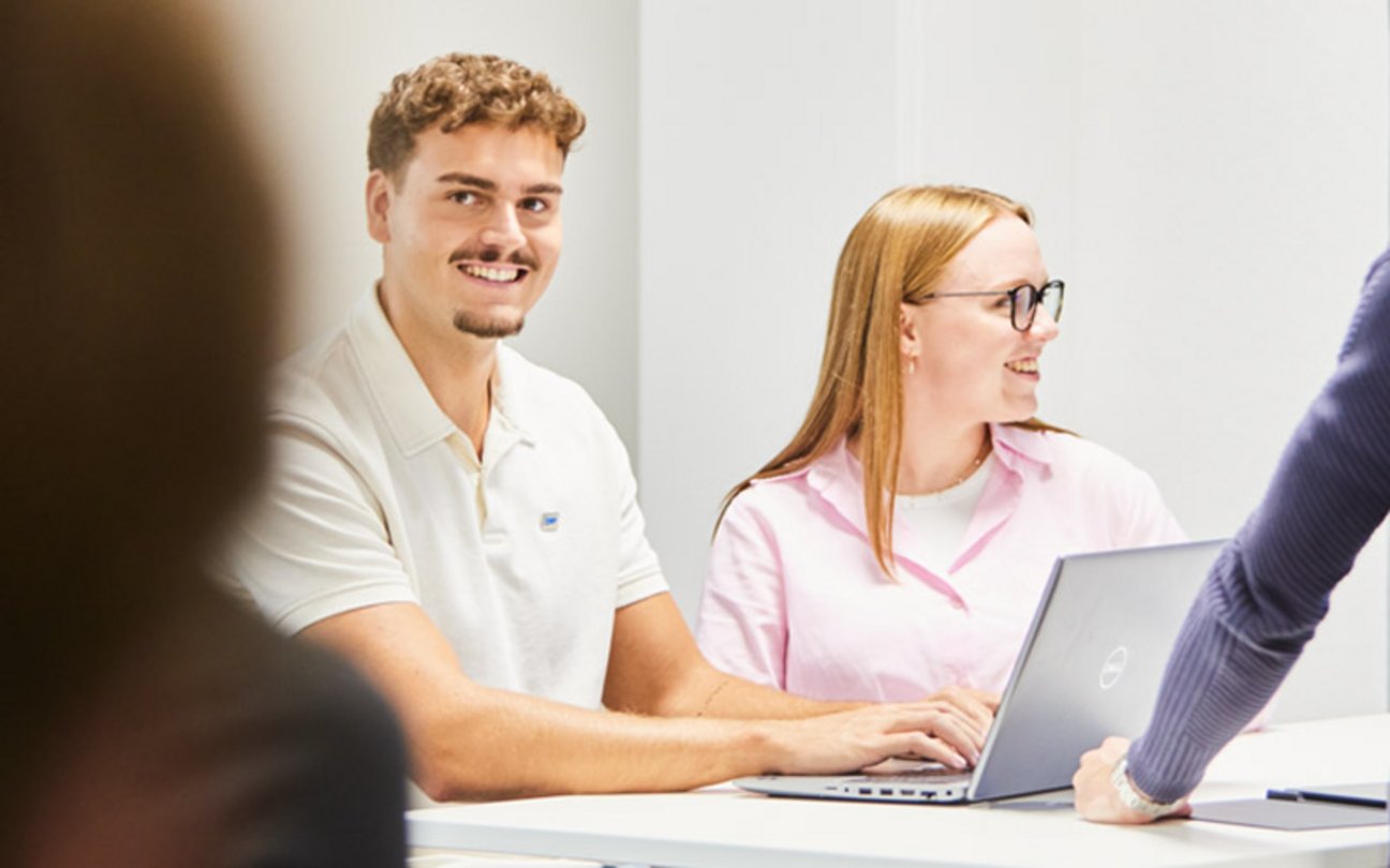 Das Bild zeigt einen jungen Mann an einem Laptop, der in die Kamera lächelt sowie neben ihm eine junge Frau, die in Richtung rechten Bildrand lächelt.