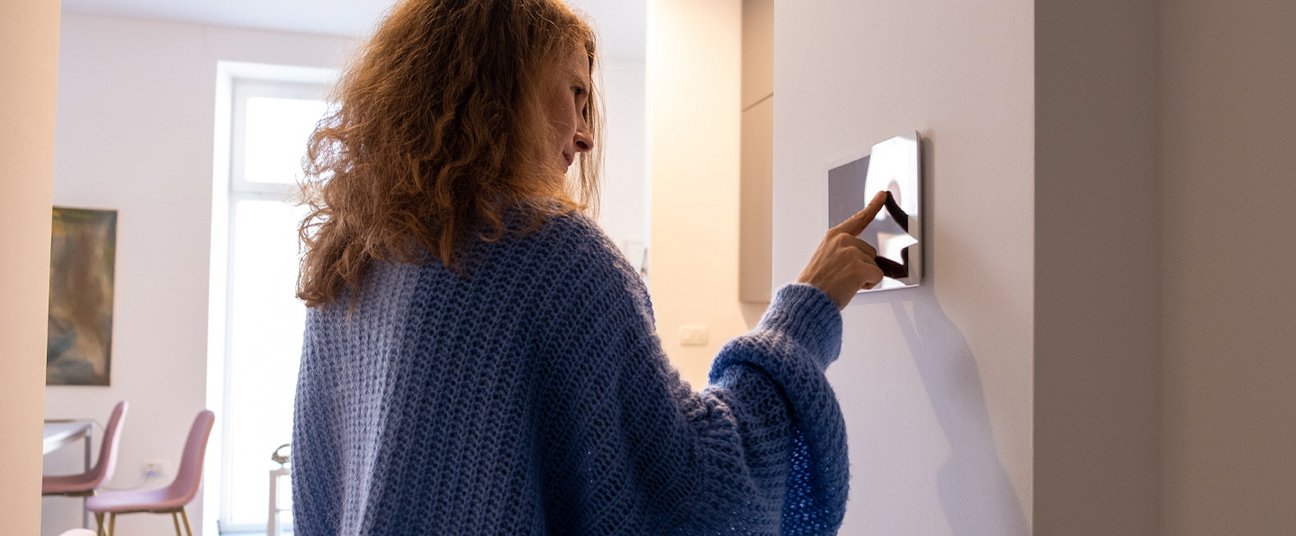 Frau mit lockigem Haar in einem blauen Strickpullover tippt auf ein smartes Wandbedienfeld in einem modernen, hellen Raum.
