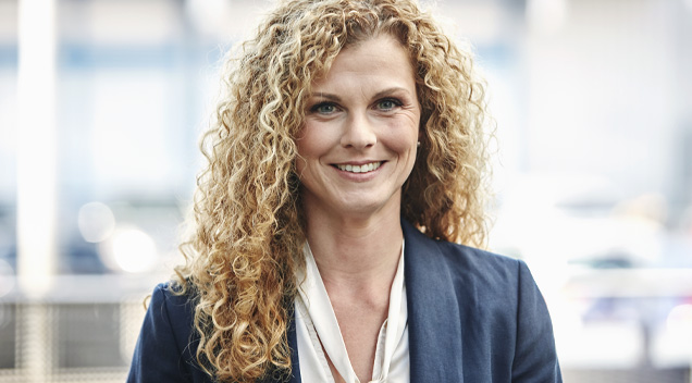 Geschäftsporträt einer Frau mit blonden Locken in Bluse und Blazer vor verschwommenem Hintergrund.