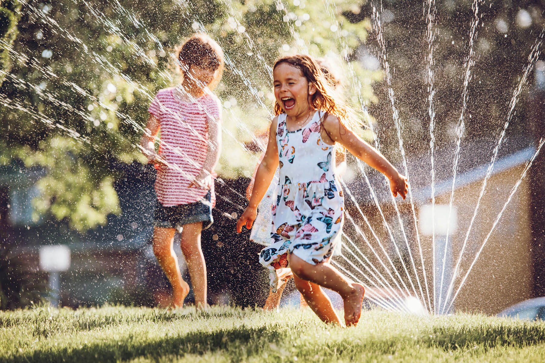 Zwei lachende Kinder spielen an einem heißen Sommertag im Wasserstrahl eines Rasensprengers